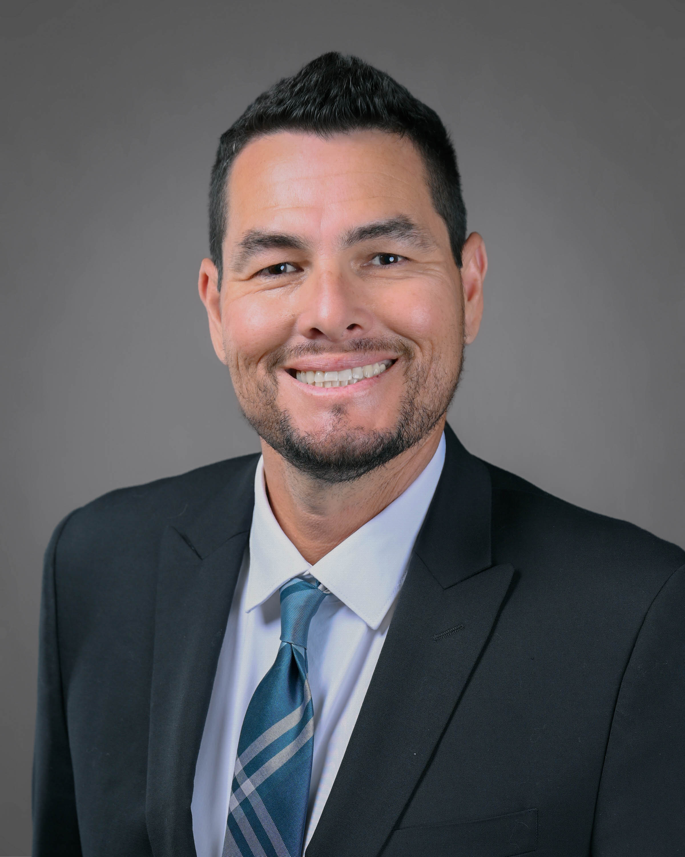 Smiling man with dark hair and short beard in a dark suit with a turquoise tie